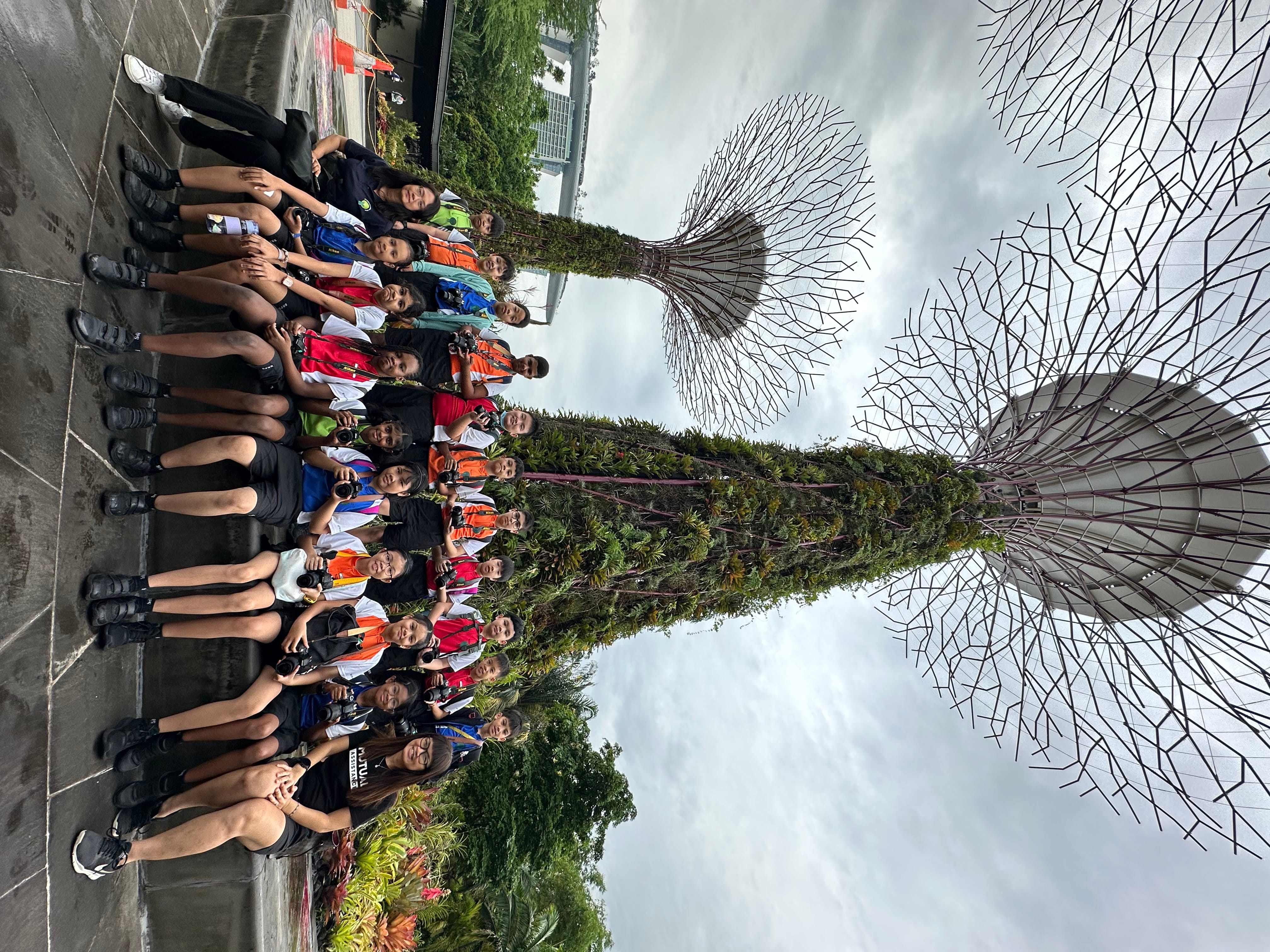 Group of teens sitting on a ledge posing for a photo, with tall metal tree towers behind them.